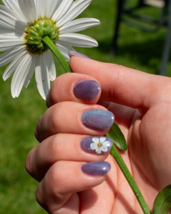 Close outdoor view of one hand holding a daisy, lilac-blue cat-eye nails with a small white daisy accent.