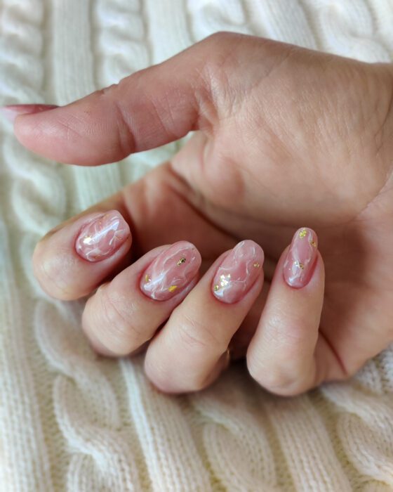 Medium oval nails in sheer pink with white marble veining and scattered gold foil, photographed on a cream knit.