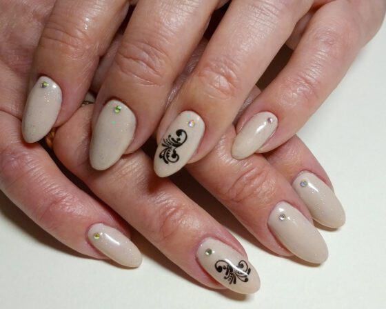 Medium oval nails in shimmering beige, two with a black baroque stamped motif and the others each set with a single crystal.