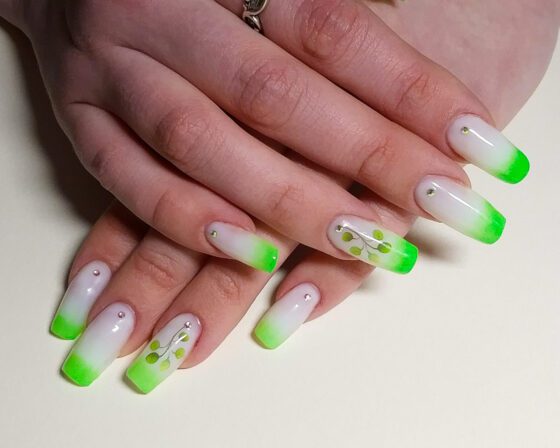 Long square extensions in milky white with neon green ombre tips, two nails carrying a leafy sprig, small crystals at the cuticle.