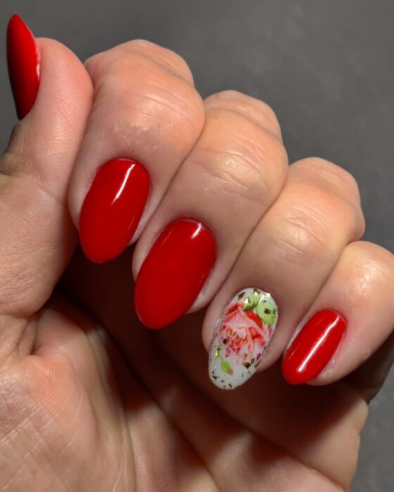 Medium almond nails in flat red gel, with one white accent nail carrying a floral slider and gold reflective flecks.