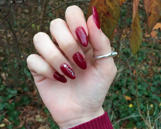 Long burgundy almond nails photographed against autumn leaves, one nail carrying silver foil at the centre.