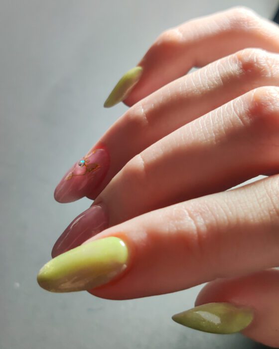 Long almond nails in translucent green and pink cat eye, with one pink nail carrying a small gold winged heart and a crystal.