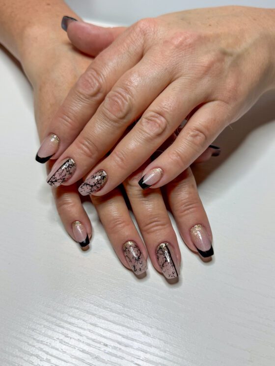 Coffin nails in nude with black French tips, gold glitter at the cuticle and a black cracked-web pattern on two nails per hand.