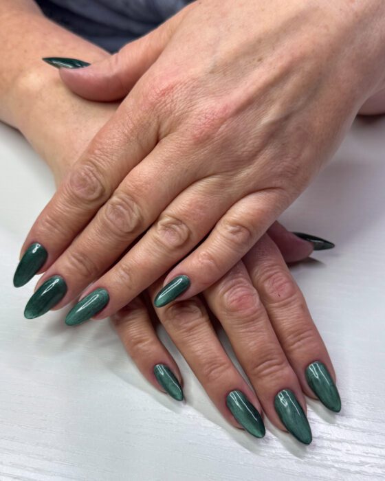 Two hands with long almond nails in deep forest green cat-eye gel, each nail showing a single band of light down its length.