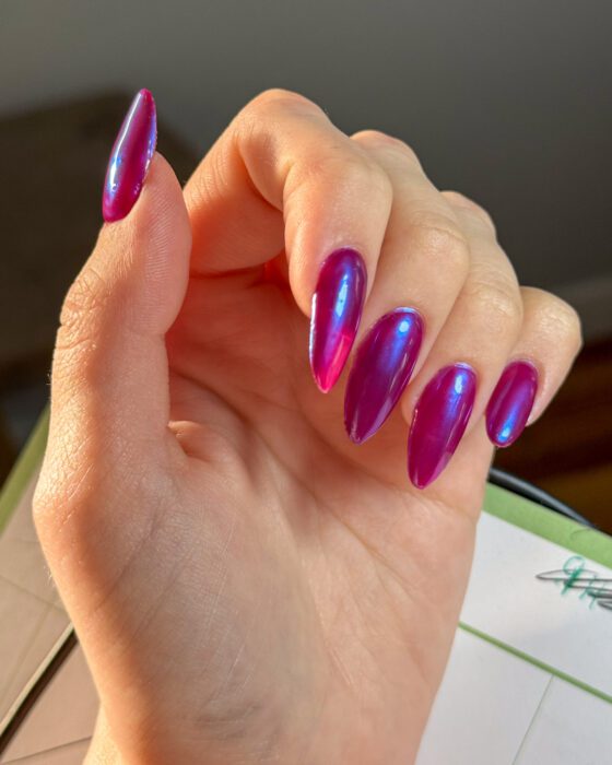 One hand with long almond nails in wine purple under a blue chrome finish, the colour shifting toward blue where the light catches it.