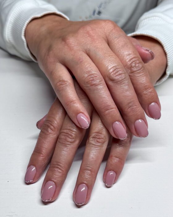Both hands resting one over the other, short oval nails in dusty nude pink with a narrow gold glitter line along each free edge.