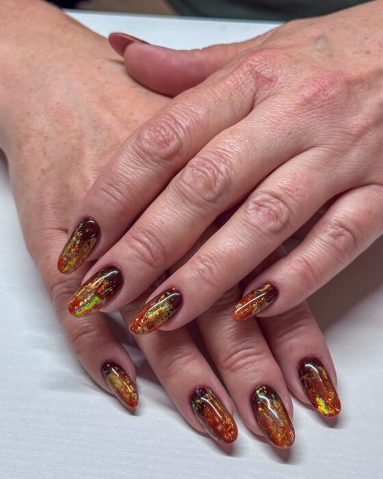 Both hands with medium oval nails in burgundy, amber, orange and gold, with iridescent foil fragments.