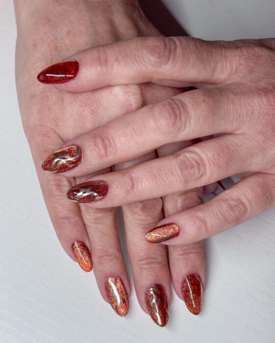 Both hands showing rust-red cat-eye nails and flowing metallic-gold line art on the deeper burgundy accent nails.