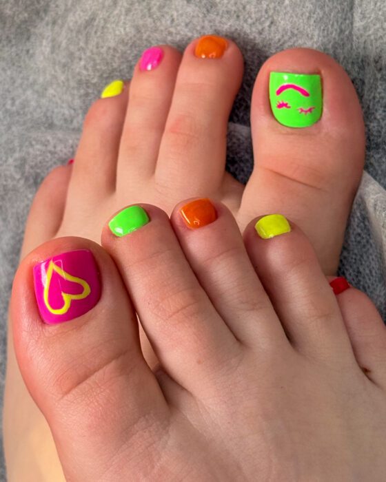 Neon pedicure in pink, green, orange and yellow, with a hand-painted yellow heart and a smiling face with lashes on the big toes.