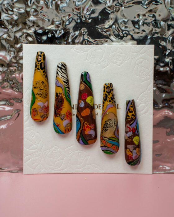 Five detached nail-art sample tips with warm orange, yellow, brown and black African-inspired motifs, including animal print and figurative details.