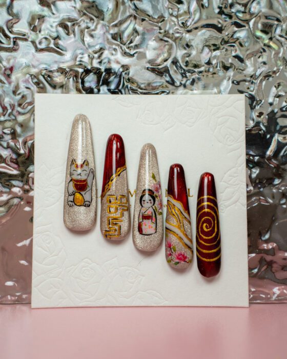 Five detached Japanese-inspired nail-art sample tips in champagne, burgundy and gold with a lucky-cat figure, a woman in traditional dress, blossoms and geometric gold line work.