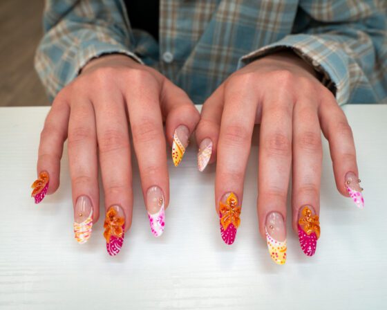 Both hands flat on a white table: extra-long almond extensions with raised orange flowers, hot-pink tips and dotted patterns.