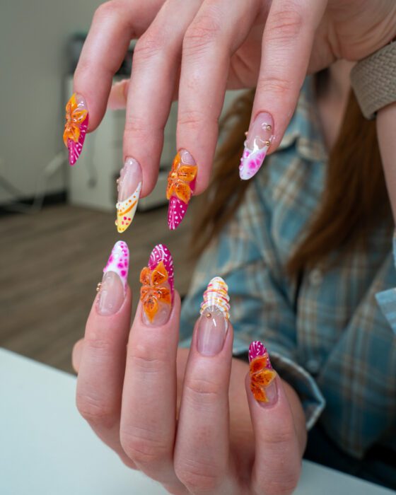 Fingers spread to show every nail: sculpted lilies, dotted pink tips, yellow striping and gold beads.