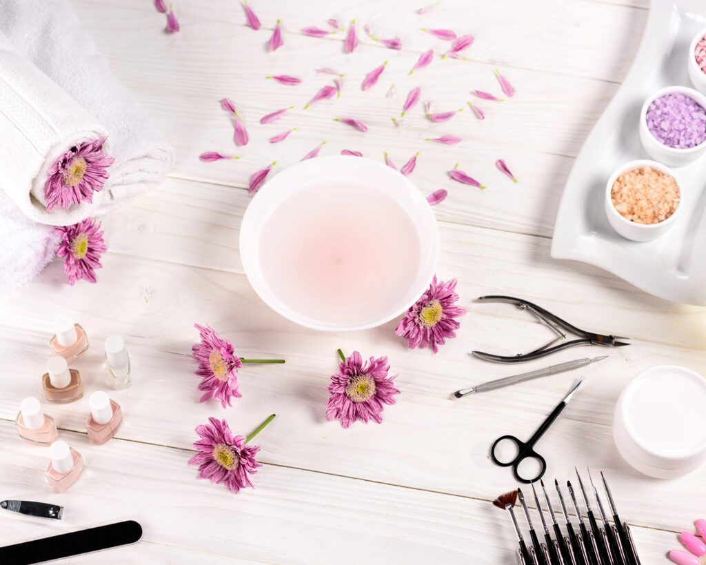 Flat lay of a manicure station with a soaking bowl, nude polishes, cuticle tools, brushes, bath salts and pink petals