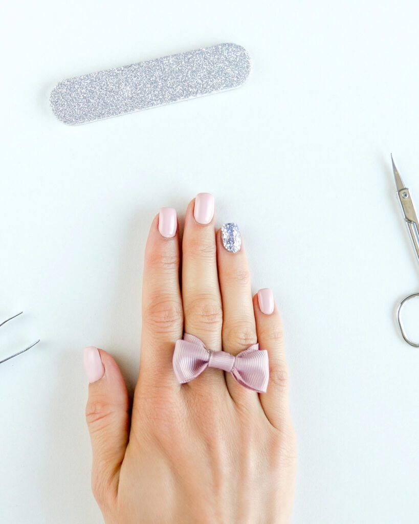 Hand with pale pink nails and one silver glitter accent, beside a glitter file and cuticle scissors