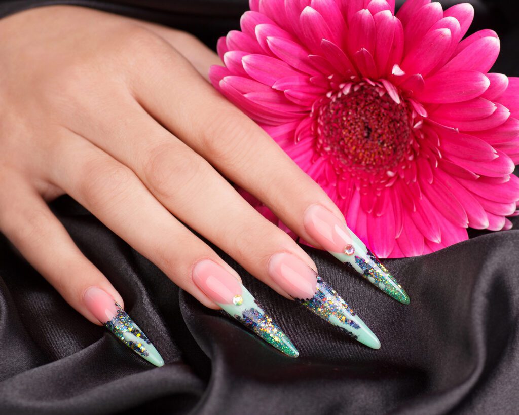 Long stiletto nails in nude and mint with glitter and gem detailing, resting on black satin next to a pink gerbera