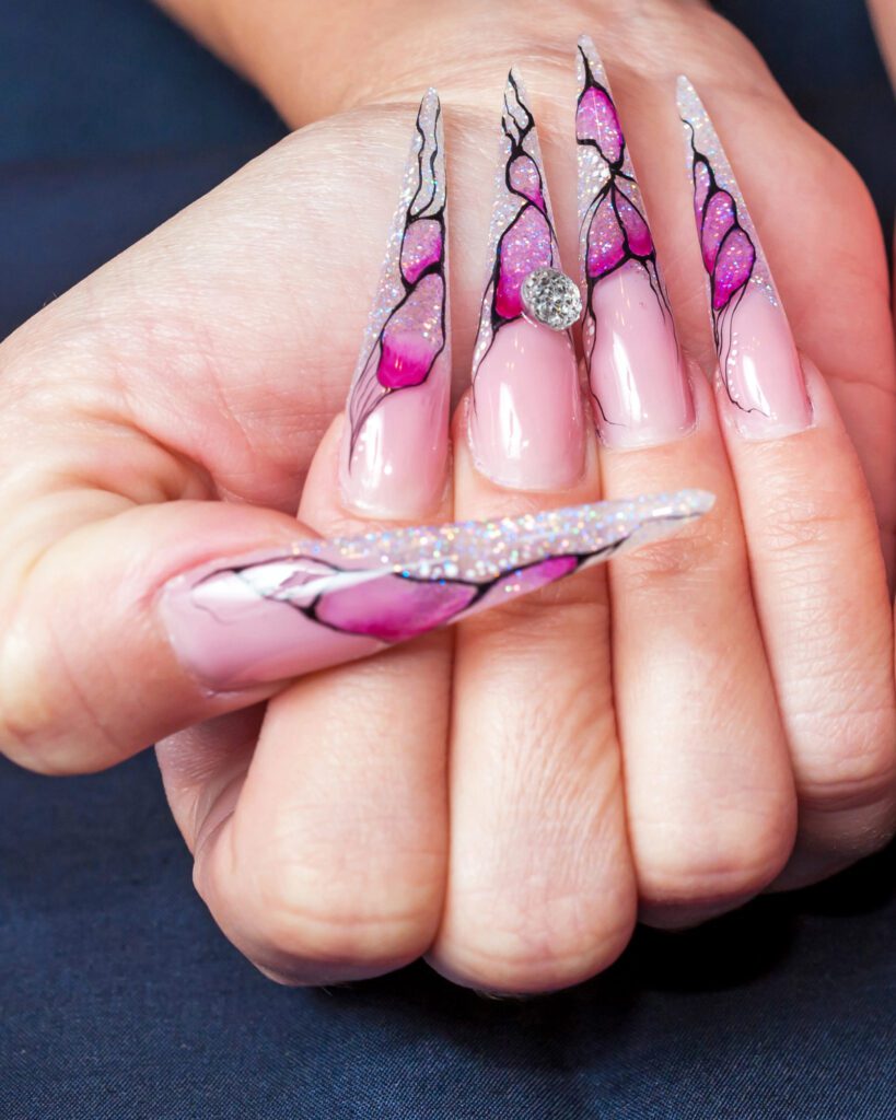 Long stiletto nails with hand-painted pink stained-glass wings, glitter tips and a crystal bead
