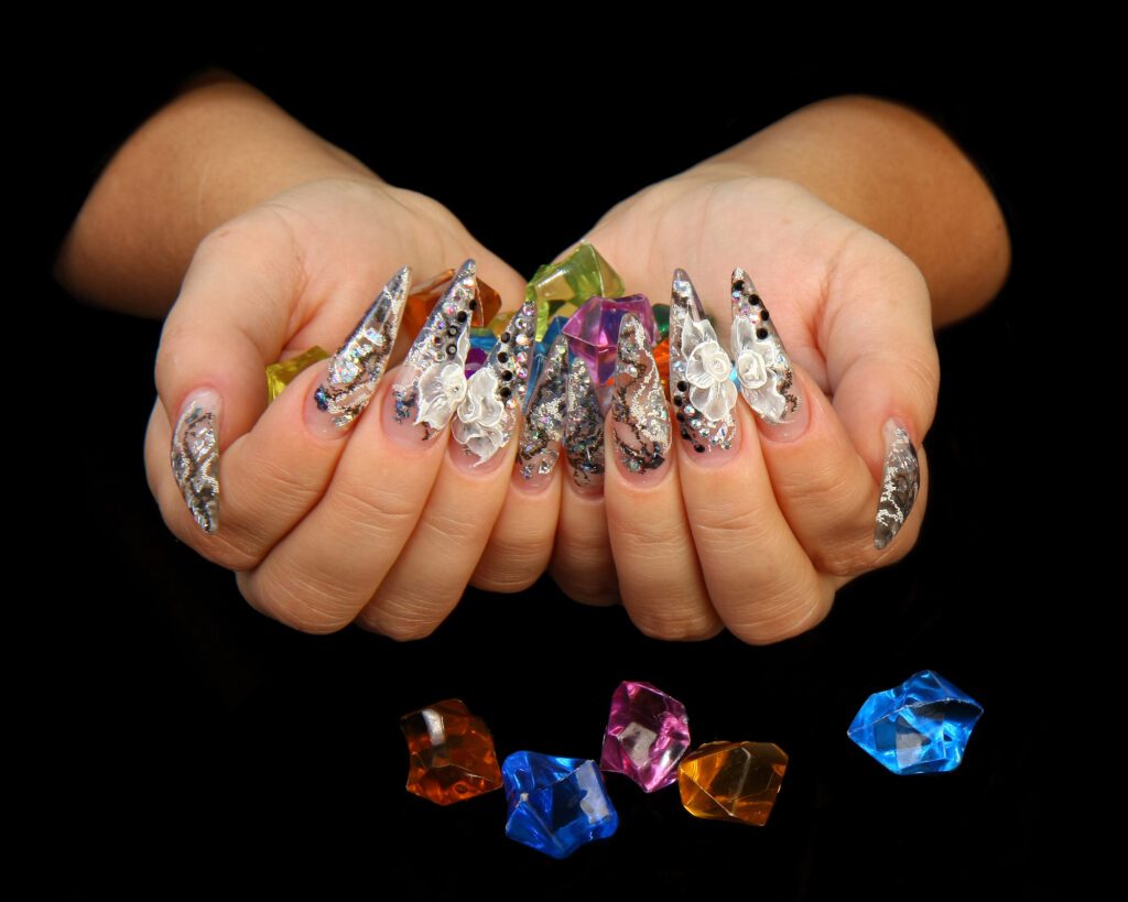 Cupped hands against a black background showing long nails in multicoloured foil art, with loose gemstones below