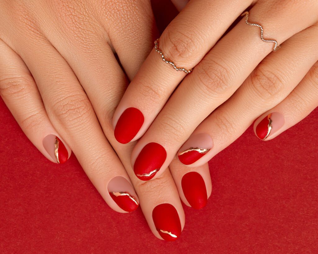 Two hands with bright red nails and thin gold accents against a red background, wearing fine gold rings