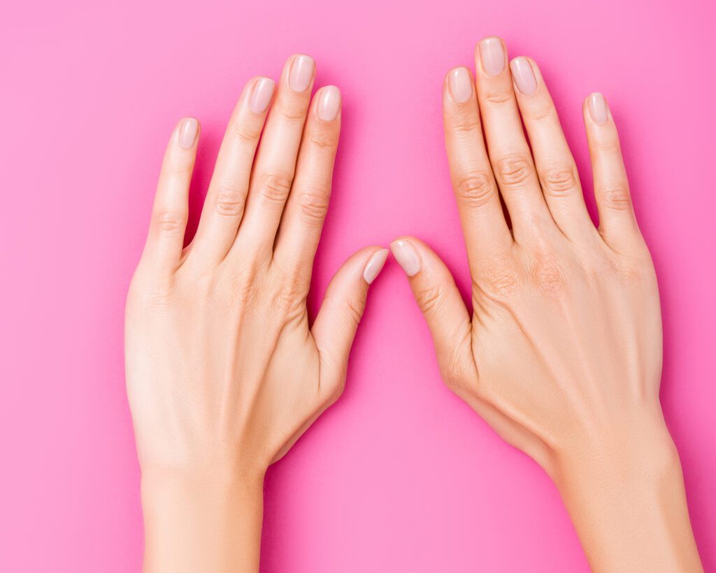 Both hands laid flat on a pink background showing short oval nails in sheer nude polish