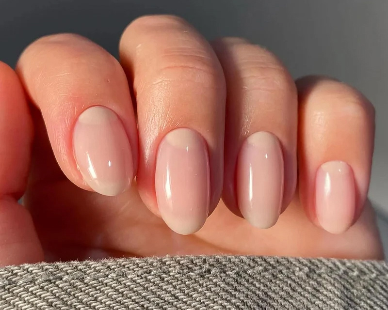 Close view of one hand with oval nails in a glossy natural pink, resting on woven grey fabric