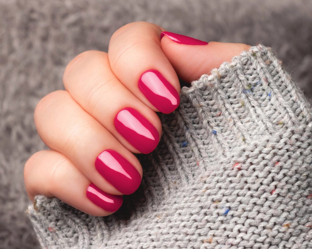 Short oval nails in glossy raspberry-pink gel polish against a grey knitted sweater