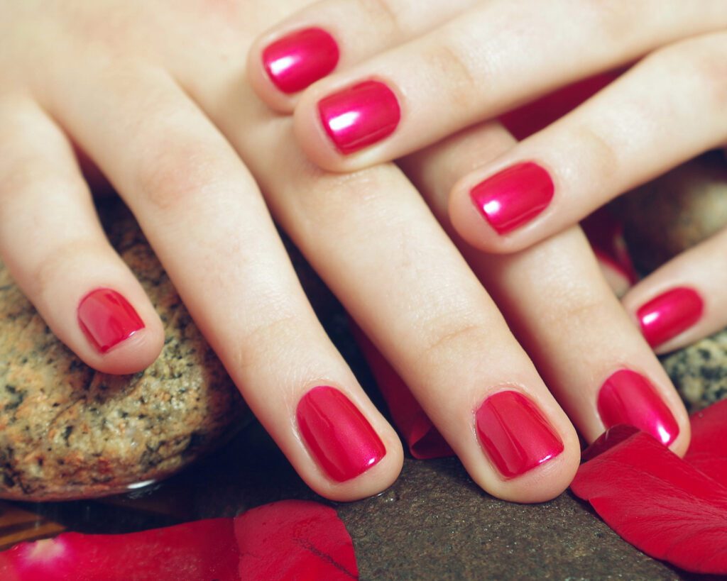 Two hands with squared nails in a deep raspberry gloss, resting on dark stone with red petals