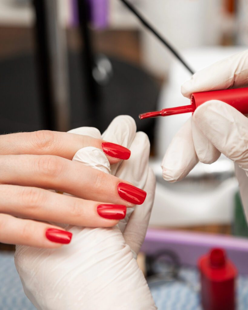 A gloved technician applying red polish to a client's nail with a brush