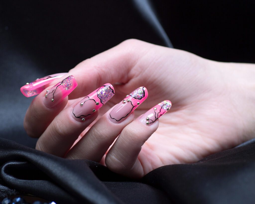 An open palm with long coffin nails in pink and clear, decorated with fine line art and glitter, on black satin