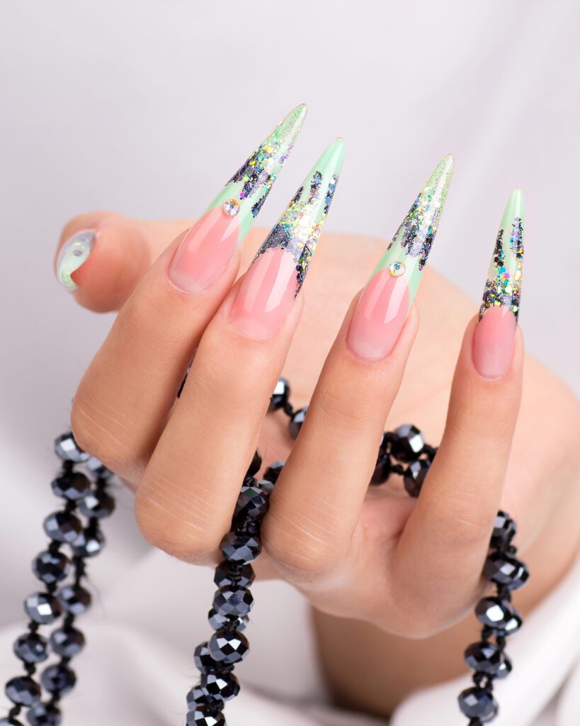 A hand with long stiletto nails in a pale green glitter gradient, resting beside a dark beaded necklace