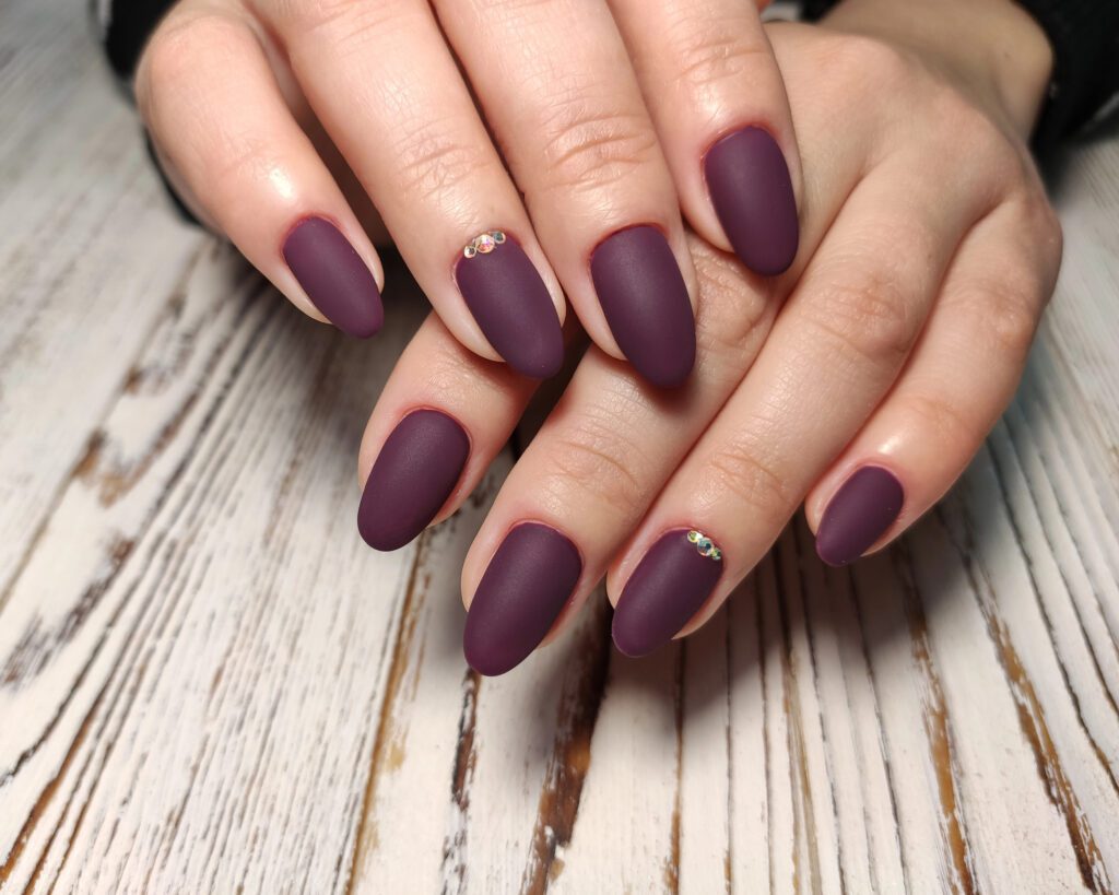 Two hands on weathered white wood with matte plum almond nails and a single rhinestone accent