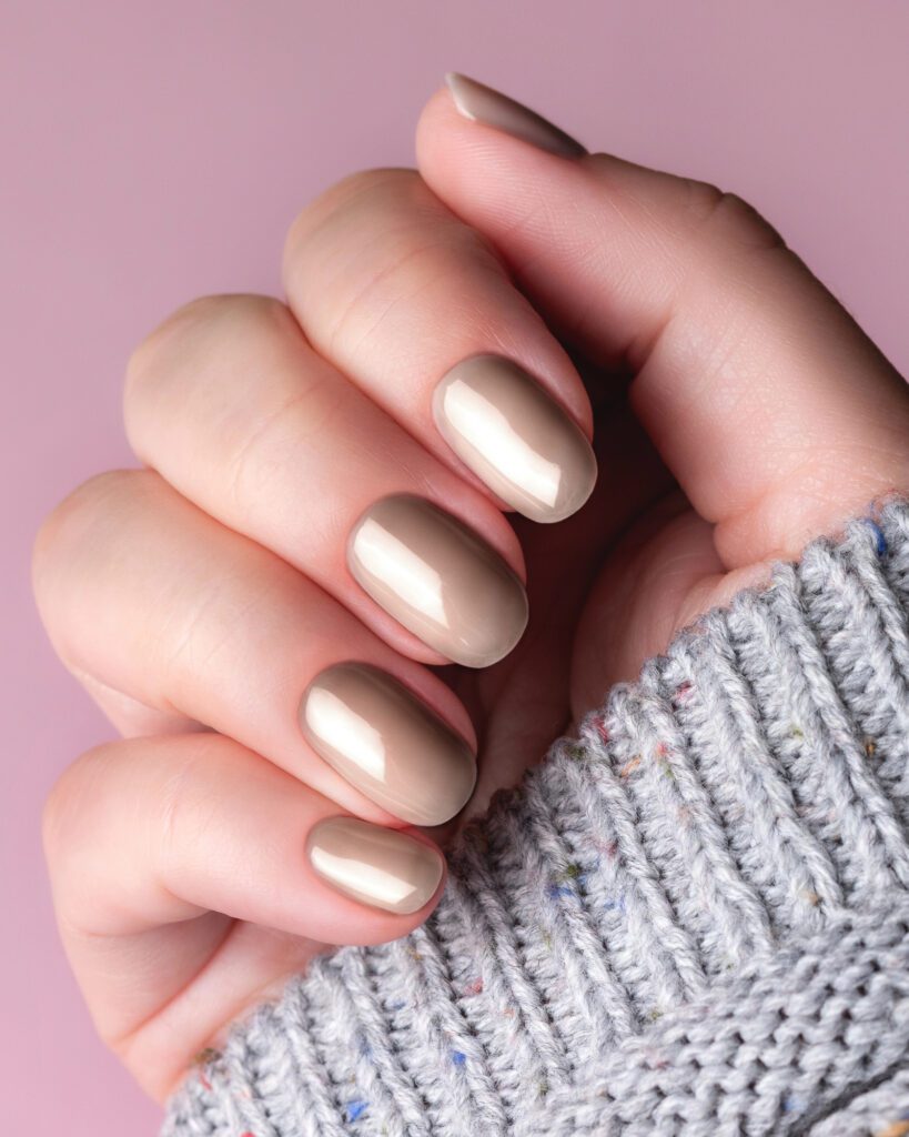 Short oval nails in glossy taupe gel against a grey knit sleeve and lilac background