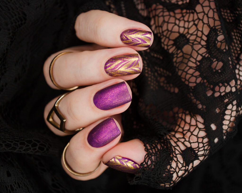 A hand against black lace with nails in purple shimmer and gold line art, wearing wide gold rings