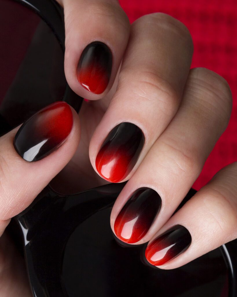 A hand with almond nails in a black-to-red gradient, resting on a polish bottle against a red background