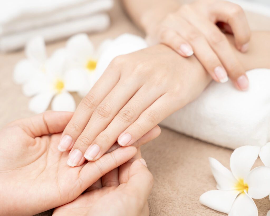One hand resting in another on a towel beside white frangipani flowers, nails in a soft natural finish