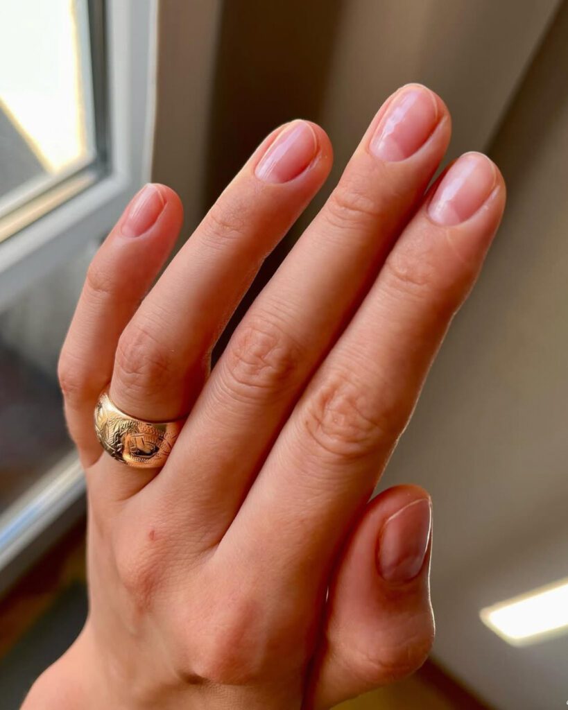A hand with short rounded nails in a bare natural finish, wearing a patterned gold ring, by a window