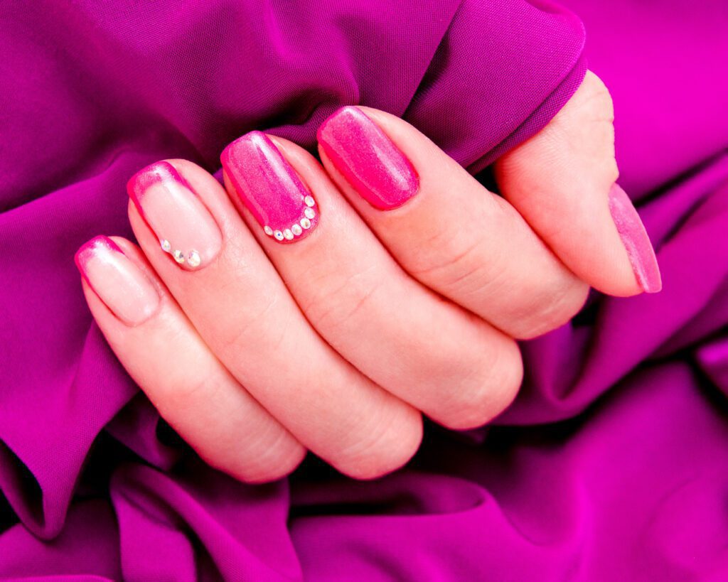 A hand against magenta fabric with pink ombre nails and small rhinestone accents