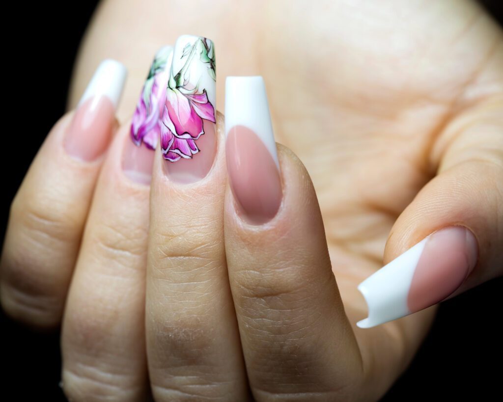 A hand against black with long coffin nails in white and nude, one painted with a pink and green floral design