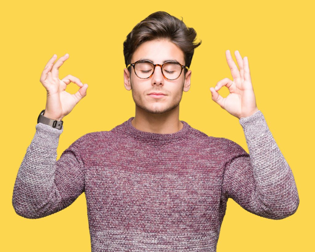 A man in a grey sweater and glasses against a yellow background, eyes closed, making a calm gesture with both hands