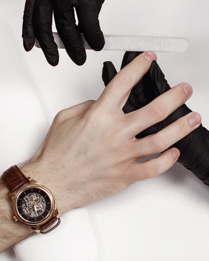 A technician in black gloves filing a man's nails, a wristwatch on his other hand