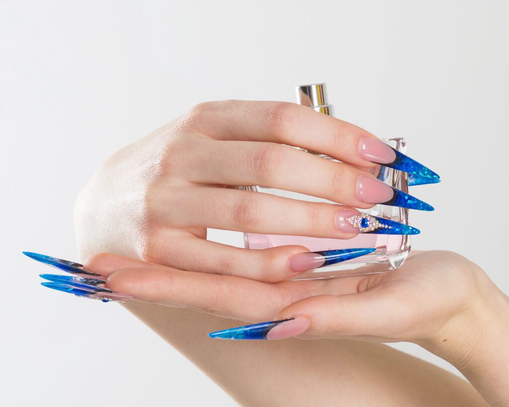 Hands with very long blue and clear stiletto nails resting against a glass perfume bottle