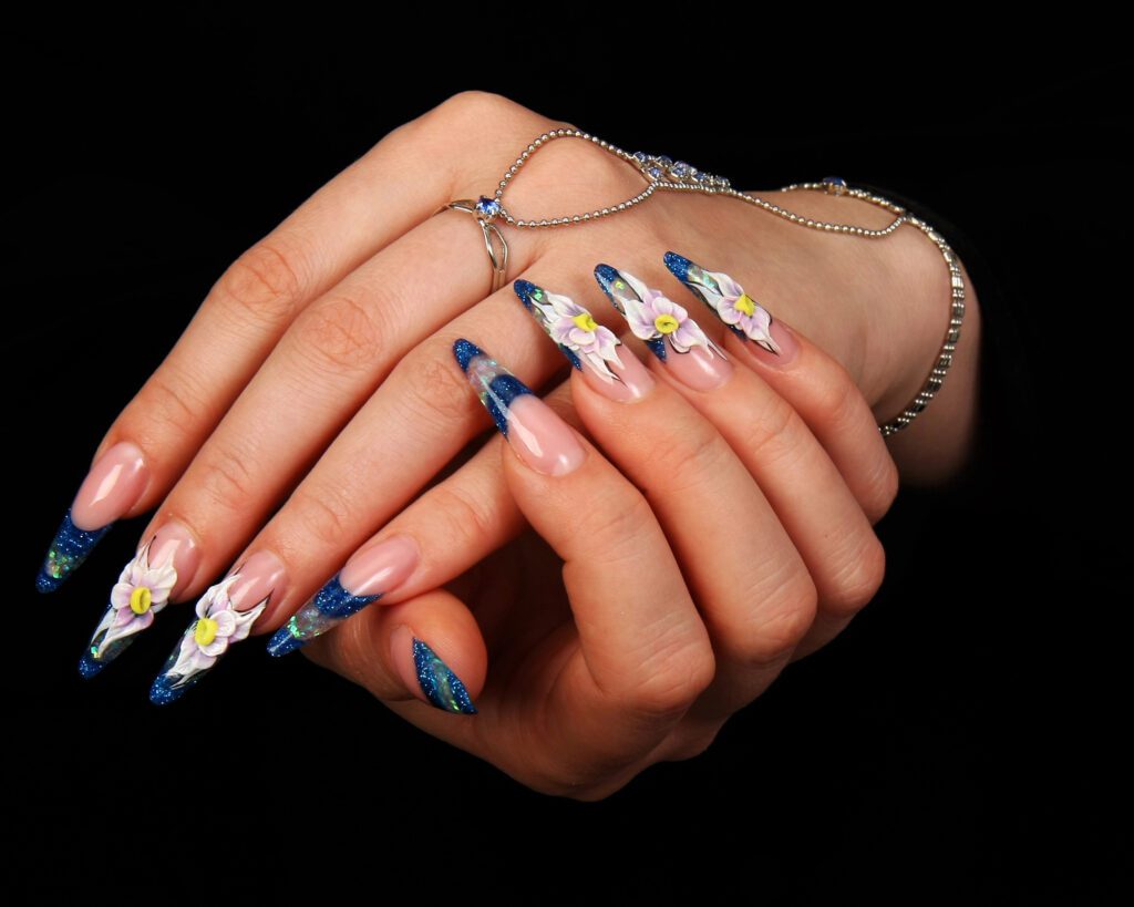 Hands against a black background with long blue glitter nails painted with white daisies, wearing a fine chain