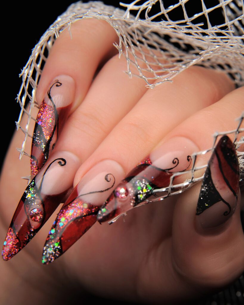 Long stiletto extensions in red and black glitter with painted swirls and crystals, behind silver mesh