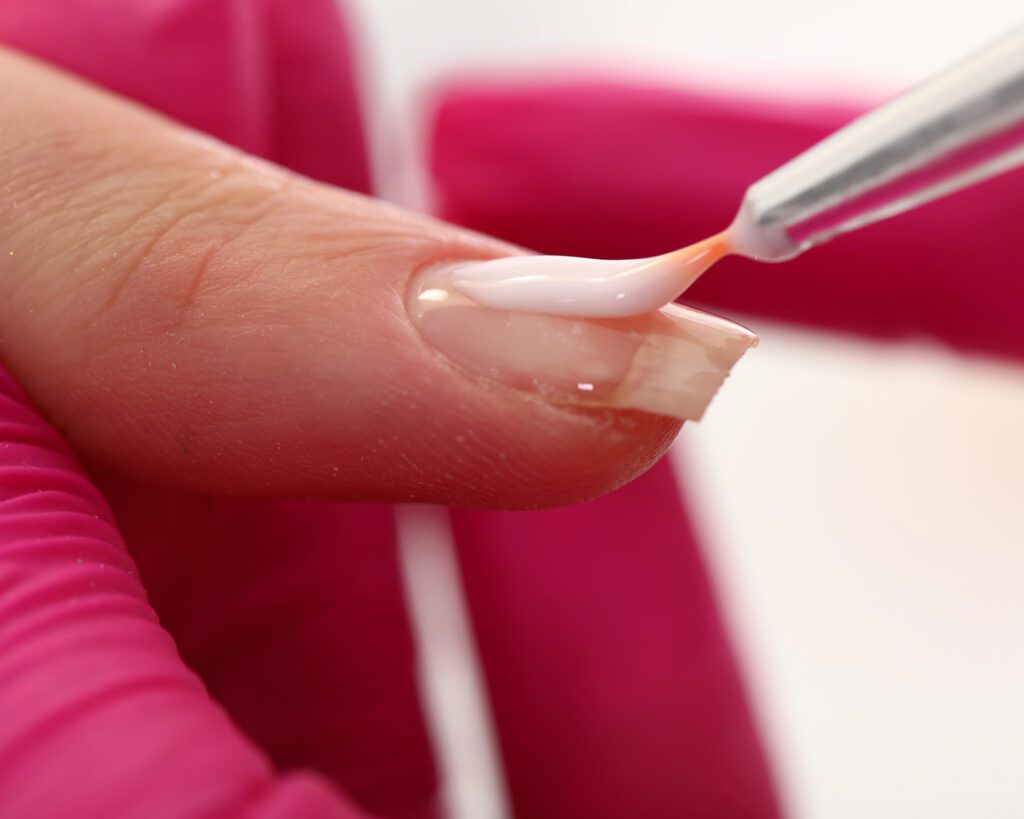 A brush applying clear gel to a single nail on a form, against a pink background