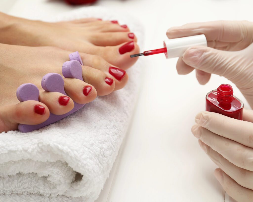 Red polish being applied to toenails with purple foam toe separators, a bottle open alongside