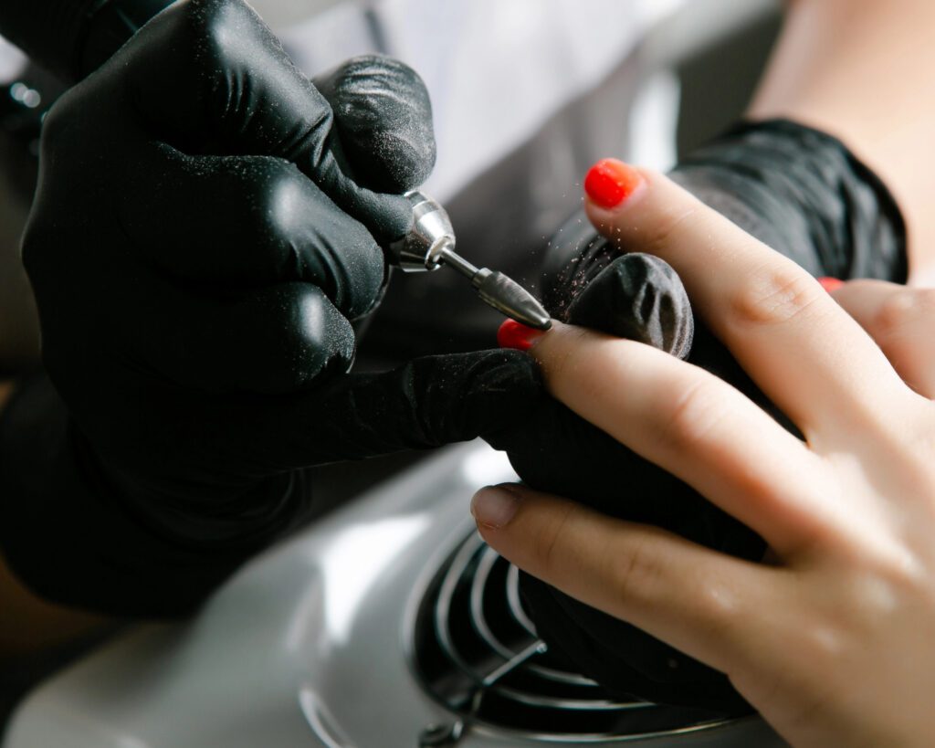 A technician in black gloves working along the nail edge with a rotary tool, one red nail visible