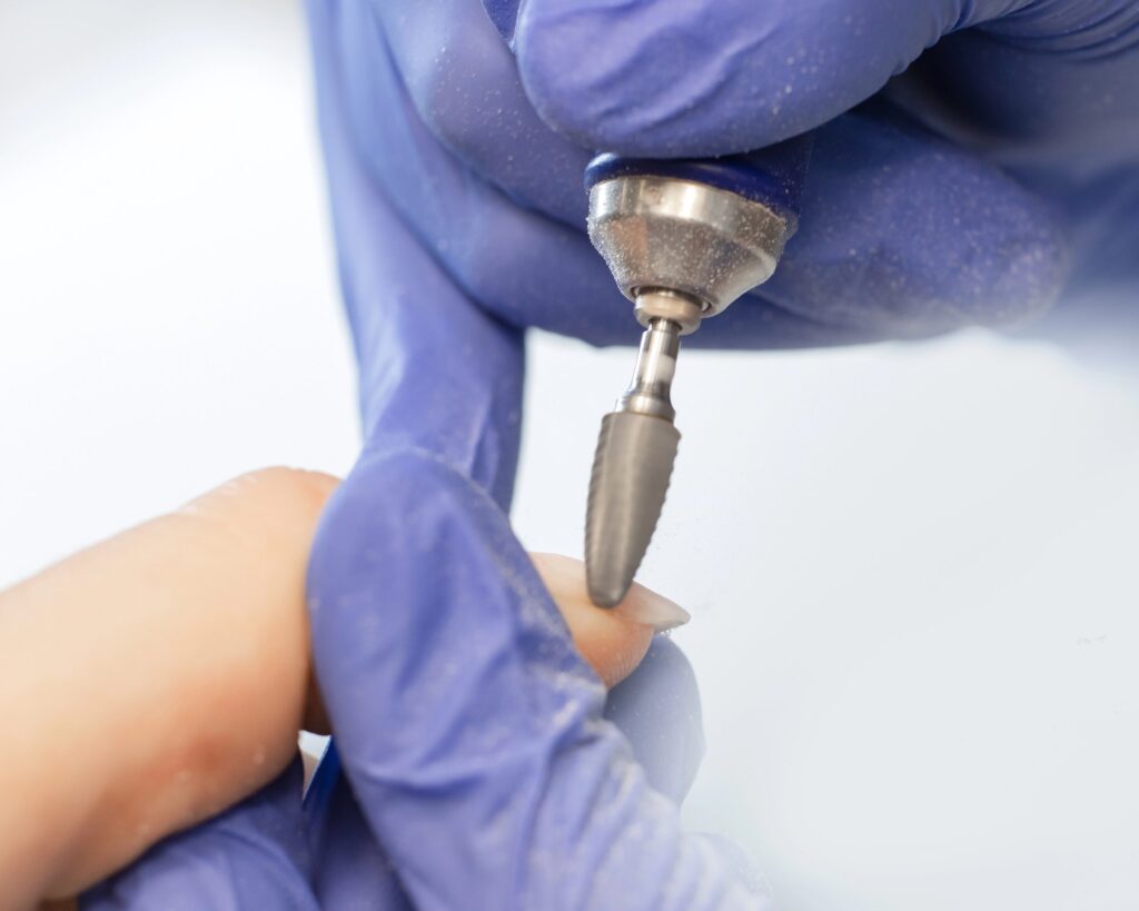 A technician in purple gloves shaping a client's fingernail with a cone-shaped rotary bit