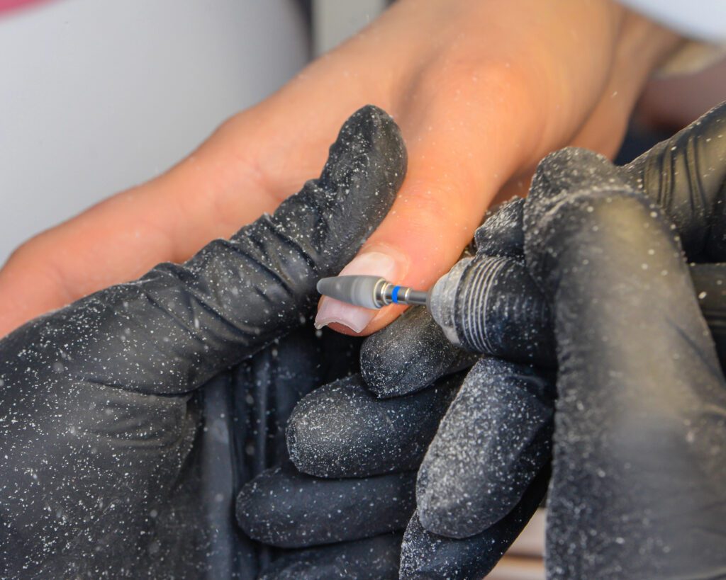 Black gloved hands using a narrow rotary bit along a nail, with visible filing dust