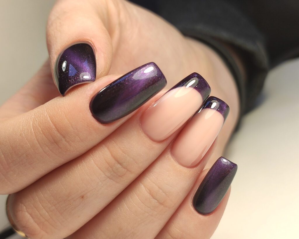 A hand with square nails in nude and purple cat-eye polish against a dark background